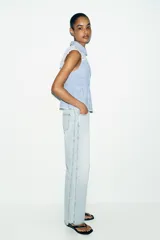 Blusa sin mangas con estampado de cuadros vichy en color azul y blanco, con cuello camisero y detalle de encaje floral en la parte superior delantera y trasera. Presenta cierre frontal con botones y un ligero volante en el ruedo.