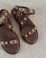 Sandalias planas de cuero negro con tiras decoradas con tachas metálicas doradas. Presentan una tira frontal sobre los dedos y una tira ajustable con hebilla alrededor del tobillo.