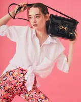 Cartera negra de cuero charolado con solapa y cierre de broche. Presenta un detalle metálico dorado en forma de freno de caballo en la parte frontal y una correa ajustable.