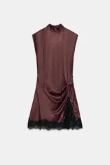 Vestido corto de cuello subido y diseño sin mangas, confeccionado en tejido satinado. Presenta un detalle de fruncido lateral con abertura frontal y aplicación de encaje negro en el ruedo.