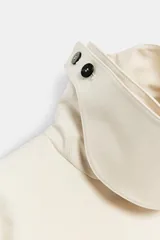 Trench corto color marfil de algodón, con cuello camisero, solapa cortaventos, mangas largas amplias con puños abotonados y cierre cruzado con un botón visible.