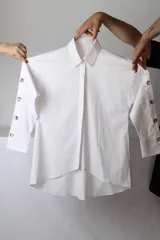 Camisa de lino y algodón color blanco, de corte oversize, con mangas largas abotonadas a lo largo del antebrazo y dobladillo asimétrico más largo en la parte trasera.