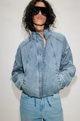 Cazadora bomber de denim con cuello subido, manga larga y cierre frontal con cremallera y botones a presión ocultos. Presenta costuras verticales decorativas y bajo con ajuste elástico.
