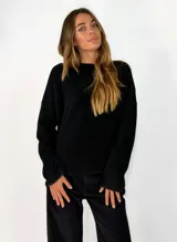 Sweater de punto negro con cuello redondo, mangas largas y diseño holgado. Presenta un detalle de terminación desflecada en el ruedo y los puños.