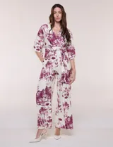 Enterito largo de calce fluido con escote en V cruzado, mangas 3/4 anchas, cintura elastizada con lazo ajustable y pantalón wide leg con bolsillos laterales. Estampado tipo toile de Jouy en tonos violeta sobre fondo blanco.