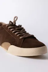 Championes urbanos de gamuza color marrón, con suela de goma en tono crema y cordones jaspeados. Presenta un refuerzo en la puntera y un detalle en cuero beige en el talón.