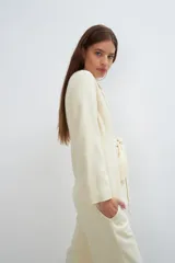 Chaqueta corta color beige, entallada, con escote en V, manga larga y bolsillos escondidos. Cierre frontal con lazo fijo. Confeccionada en lana fina sin teñir. Diseño exclusivo de Don Baez.