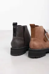 Botines de cuero vacuno color suela con textura envejecida, suela track negra con pespunte en contraste, solapa cruzada con gran hebilla metálica plateada y cierre lateral interior.