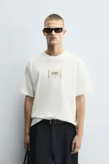 Camiseta boxy fit de color blanco con cuello redondo y mangas cortas. Presenta un parche rectangular en el centro del pecho con texto impreso y costuras decorativas a contraste en los laterales.
