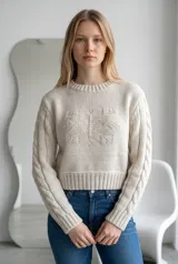Sweater de punto color verde, con cuello redondo, diseño de trenzas en las mangas y detalle de relieve en el frente. Presenta un corte corto con puños y ruedo acanalados.
