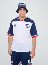 Remera deportiva de manga corta Umbro del Club Nacional de Football, modelo Match. Diseño blanco en el cuerpo con mangas, hombros y detalles en cuello en azul, con ribetes en rojo y blanco.