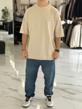 Remera oversize color beige, con mangas cortas y cuello redondo. Presenta un estampado tipográfico en la espalda con la frase "There are no facts, only interpretations" y otros detalles de diseño.