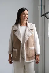 Chaleco de gamuza color beige con interior y detalles de corderito color crudo. Presenta solapas amplias, bolsillos frontales aplicados y diseño sin mangas.