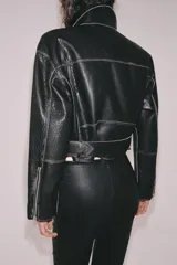 Chaqueta tipo biker de piel negra con solapas, cierre frontal asimétrico con cremallera, bolsillos con cierre y cinturón ajustable con hebilla metálica en la cintura.
