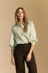 Camisa blanca de manga larga con cuello y botones frontales. Presenta detalles de volados o puntilla en los hombros y mangas abullonadas con puños anchos. La tela parece tener un sutil tejido a rayas verticales.