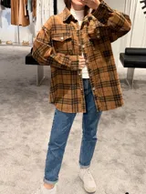 Camisa tipo sobrecamisa de corte oversize, confeccionada en tela a cuadros en tonos marrones y negros. Presenta cierre frontal con botones metálicos a presión, bolsillos frontales con solapa y mangas largas con puños abotonados.