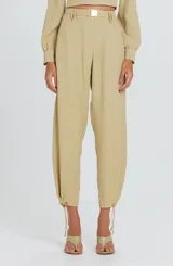 Pantalón rosado de tiro alto con cintura estructurada y hebilla metálica decorativa, diseño fluido de corte relajado con pliegues frontales y ajuste con cordón en los tobillos.