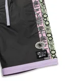 Boardshort de 17 pulgadas, color negro, con pretina elástica color lila y cordón de ajuste gris. Presenta paneles laterales con estampado gráfico en tonos rosa pálido y verde menta. Incluye un parche bordado con el logo "T.C.S.S" en la pierna izquierda y otro parche con el texto "CRITICAL SLIDE" en la derecha.