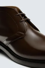 Bota de caña baja tipo chukka, confeccionada en piel con acabado brillante. Presenta cierre de cordones, horma redonda y suela a contraste con ligero tacón.