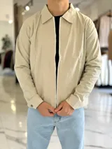 Campera de gabardina beige de corte boxy fit, con cuello camisero, cierre frontal de cremallera blanca y bolsillos laterales ribeteados.