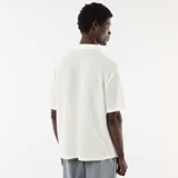 Camisa de manga corta color blanco, confeccionada en tejido de punto calado con patrón de rayas verticales sutiles, cuello abierto tipo campamento y cierre frontal de botones.
