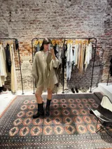 Ruana tejida a mano en lana merino color beige, con textura de punto y detalle de flecos en el ruedo. Presenta sutiles destellos brillantes integrados en el tejido.