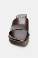 Sandalia de piel color marrón tipo zueco, con tira delantera ancha, detalle de tachas metálicas doradas en los laterales, tacón en bloque con efecto madera y puntera cuadrada.