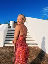 Vestido largo de crepé con estampado floral en tonos rosados, morados y blancos. Presenta escote buche con drapeado y espalda baja ajustable con tiras finas. El ruedo es irregular, más corto en el frente.