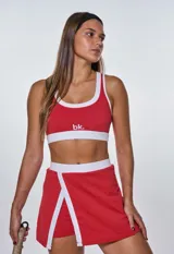 Conjunto deportivo de dos piezas compuesto por un top deportivo con escote redondo y una pollera short con abertura frontal, ambos en color rojo con detalles en blanco.