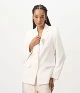 Blazer estructurado color blanco de la marca Cortelle, con solapa clásica, bolsillos frontales con solapa, mangas largas y cierre frontal de botones. Incluye un broche decorativo dorado con flecos de pedrería.