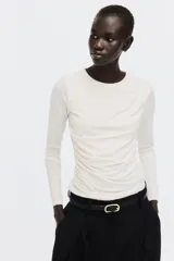 Top de punto de crepé color beige, de ajuste ceñido, con mangas largas y escote redondo. Presenta un diseño con frunces en un hombro y en un lateral que genera un efecto drapeado.