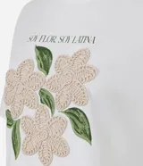 Remera de algodón color blanco, con cuello redondo y manga corta. Presenta un estampado con la frase 'Soy flor. Soy latina' y flores bordadas en relieve con textura tejida.