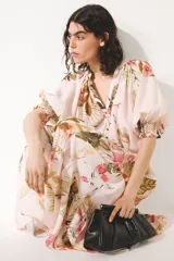 Vestido midi oversized de mezcla de viscosa, con estampado floreado tropical sobre base rosada, escote en V y botonadura frontal. Cuenta con mangas al codo raglán con puños fruncidos, cinturón fino para atar con cuentas y diseño escalonado en la falda.