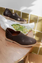 Zapatos tipo mocasín de cuero color marrón oscuro, con diseño de textura lisa en la parte delantera y textura de reptil en el talón. Cuentan con cierre trasero y suela plana de color claro.