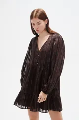 Vestido corto de manga larga con cuello en V y cierre frontal de botones. Presenta un diseño holgado con falda de volado en el ruedo y un estampado sutil de rayas verticales brillantes sobre fondo oscuro.