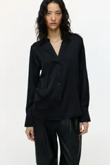 Blusa negra de tela satinada con brillo, cuello clásico, escote en V con botones frontales, mangas largas con puños sencillos y bajo redondeado.
