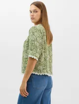 Blusa de gasa estampada con motivos botánicos en color verde, con escote redondo, abertura frontal con lazos de ajuste y mangas cortas. Presenta detalles de encaje en el escote, mangas y ruedo.