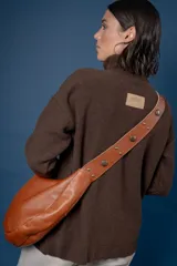 Bolso tipo bandolera de gamuza color marrón, con correa ancha ajustable decorada con tachas metálicas y cierre superior.