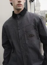 Camisa de corte boxy con estampado de cuadros pequeños en tonos oscuros. Presenta cuello camisero en color negro, cierre frontal con botones, canesú en la parte superior y un bolsillo en el pecho con etiqueta de la marca Kapsul Studio.