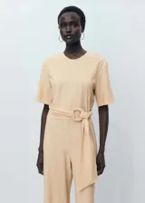 Mono largo de diseño recto, color beige, con cuello redondo, mangas cortas y cierre de botón en la parte posterior. Incluye un cinturón integrado con hebilla del mismo tejido para ajustar a la cintura.