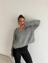 Buzo de punto grueso color gris, con cuello polo, cierre frontal parcial y capucha. Presenta un diseño de tejido acanalado con terminaciones desflecadas en el ruedo.
