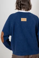 Saco de punto color beige, con cierre frontal de botones marrones y detalle de costuras decorativas oscuras en los hombros. Presenta un diseño clásico con cuello redondo.