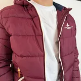 Campera tipo puffer color bordó con capucha, cierre frontal blanco y puños de tejido elástico. Presenta el logo de la marca bordado en el pecho.