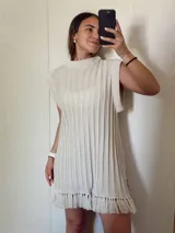 Vestido corto color beige tejido con hilo de algodón y lurex en punto acanalado vertical y flecos. Tiene corte recto, cuello redondo con una guarda ancha y hombros amplios que dan efecto de hombrera caída.