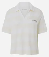 Remera polo deportiva de manga corta, confeccionada en tejido piquet con textura. Presenta un diseño a rayas horizontales en tonos crema y blanco, con cuello tipo polo y bordado con la palabra 'Athletics' en el pecho.