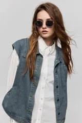 Chaleco de jean azul con sutil textura a rayas, cuello camisero, cierre frontal con botones metálicos y diseño sin mangas con hombros amplios.