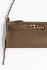 Cartera rectangular de hombro, color marrón topo, con textura de gamuza o nobuck. Presenta un cierre superior y una tira decorativa frontal con herraje metálico dorado tipo brida de caballo. Incluye correa larga ajustable para llevar al hombro.