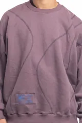 Buzo blanco de manga larga con diseño holgado, cuello redondo y costuras externas visibles en contraste. Cuenta con una etiqueta decorativa en el frente inferior.
