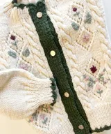 Cardigan de lana merino color marfil, con bordados florales artesanales, trenzas y rombos calados en el frente. Posee cierre frontal con botones vintage dorados y terminaciones en crochet celeste en cuello, puños, ruedo y tira de botones.