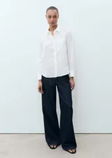 Camisa de lino blanca de corte recto con cuello camisero, manga larga con puños abotonados, cierre frontal de botones y un bolsillo de parche en el pecho.